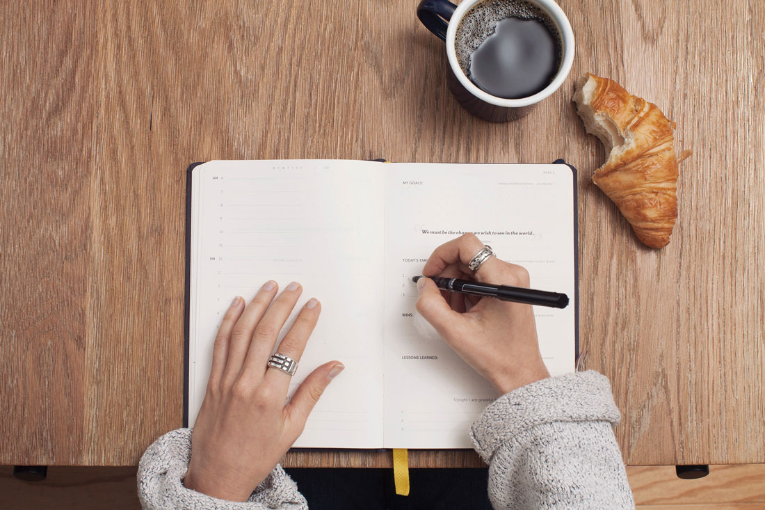 Bild zeigt Hände die in Notizbuch schreiben, mit Kaffee und Croissant
